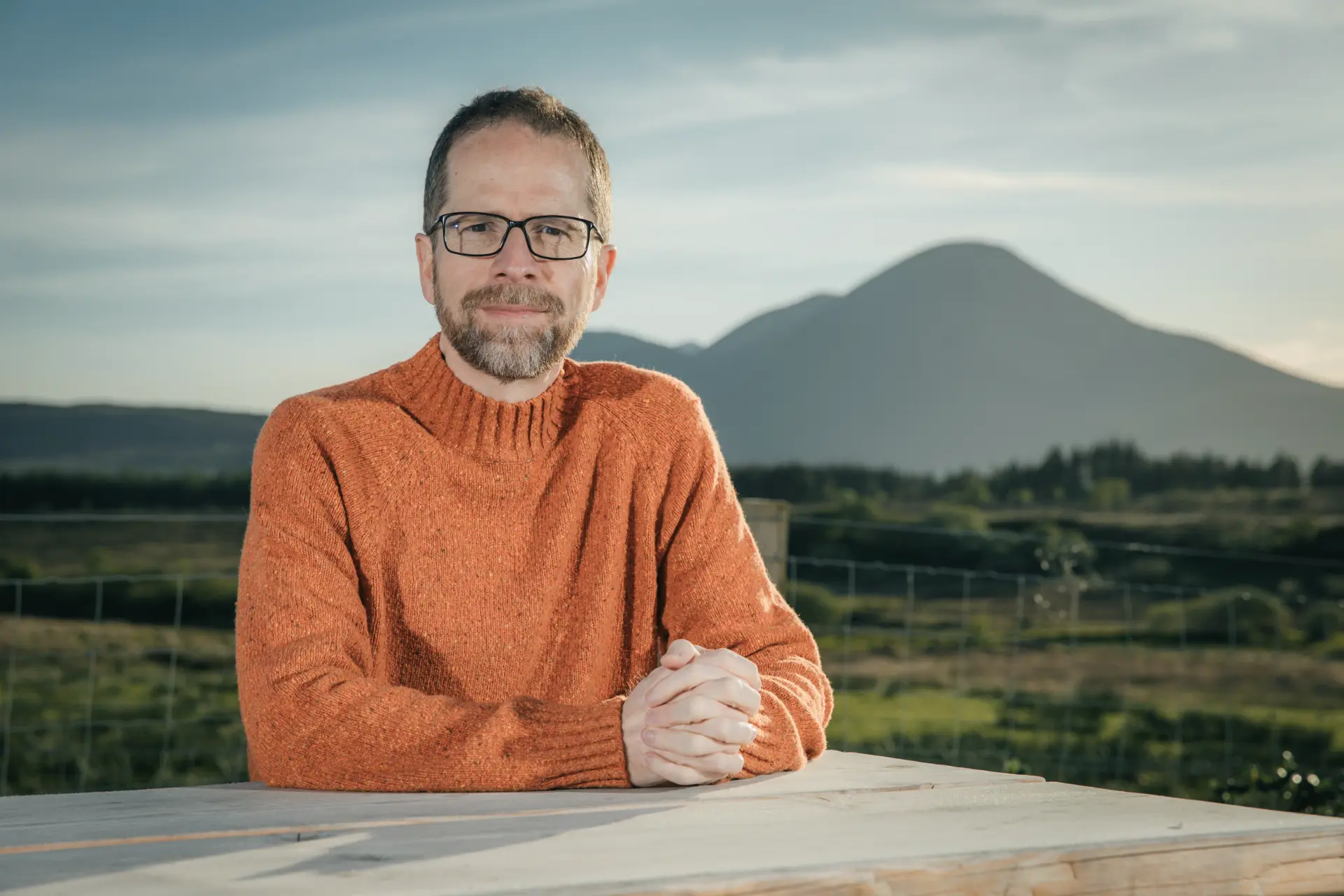 Matthew Savage seated at a table outdoors, hands folded, mountains behind him